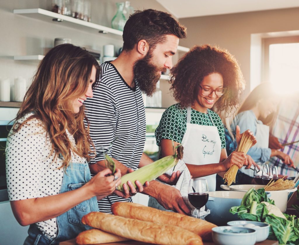 Good friends laughing and talking while preparing meals at table full of vegetables and pasta ready for cooking in kitchen